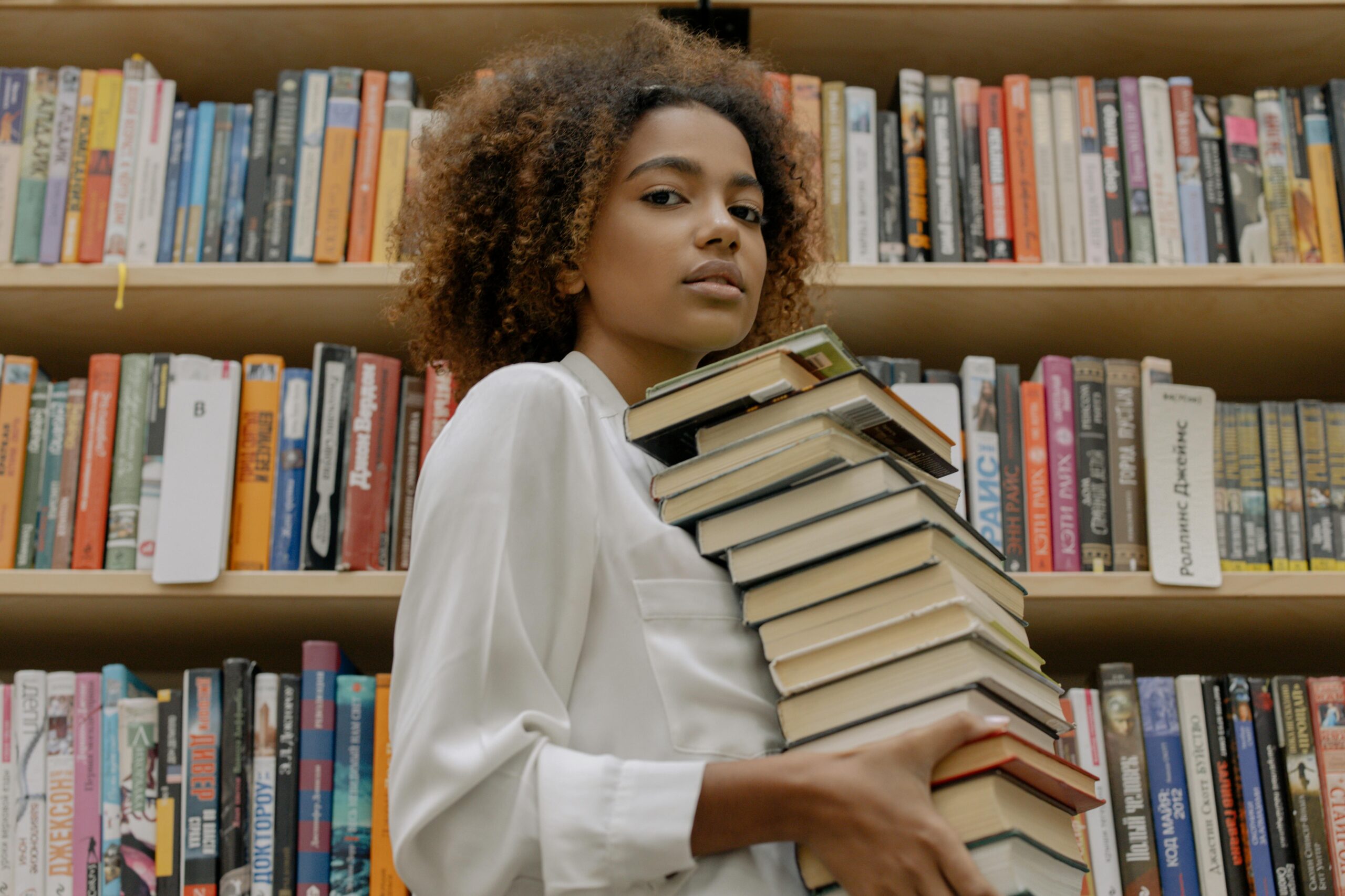 A young woman carrying a stack of books while she is walking by tall bookshelves